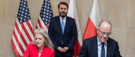 Deputy Minister of Climate and Environment Krzysztof Galos and Under Secretary of State Allison Hooker sign documents at a table during an official ceremony; both are dressed in formal attire—the woman in a red suit and the man in a business suit—while a third man in a suit stands behind them, with Polish and U.S. flags in the background.