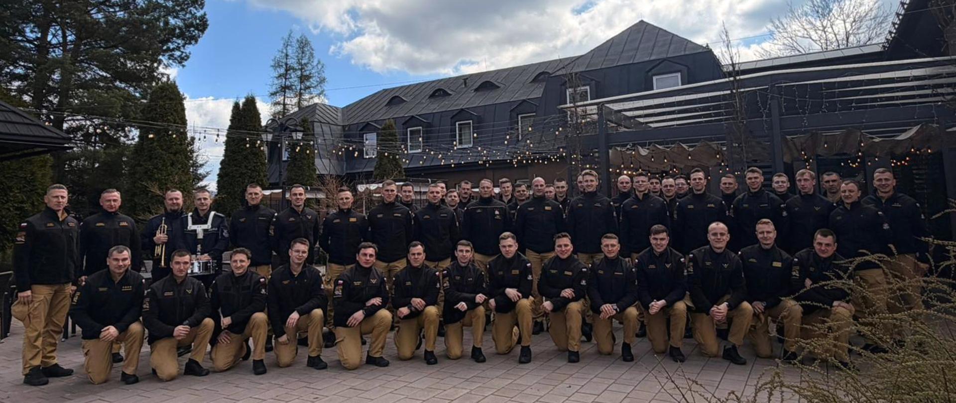 Na placu, na tle zabudowań stoi grupa strażaków w czarnych polarach służbowych i piaskowych spodniach. Rząd z tyłu stoi, rząd z przodu kuca. Strażacy zwróceni są przodem do aparatu, pozują do zdjęcia grupowego