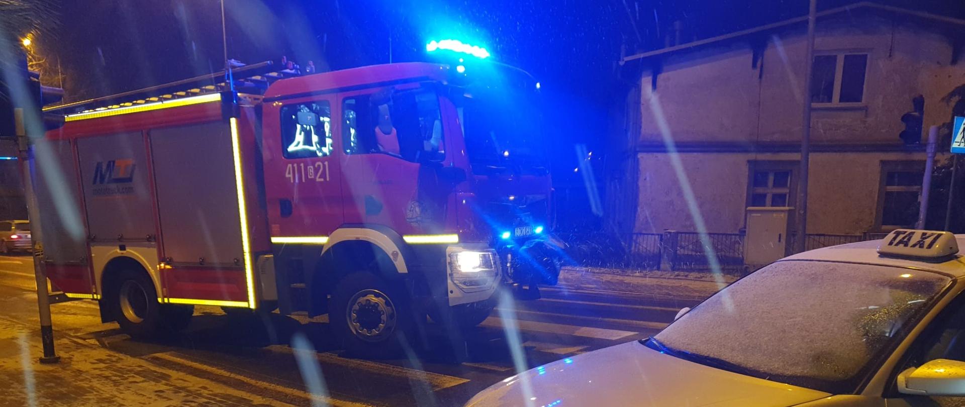 Pora nocna, na pasie ruchu stoi czerwony samochód straży pożarnej wysyłający niebieskie sygnały świetlne. Na pierwszym planie widać srebrny samochód z uszkodzonym lewym przodem. Za pojazdami widoczny budynek mieszkalny. 