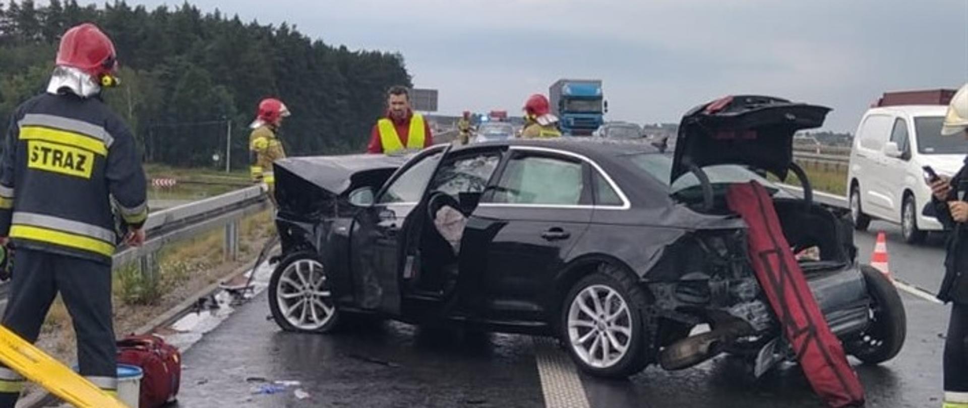 Na zdjęciu widoczny uszkodzony pojazd osobowy oraz strażacy wraz ze sprzętem ratowniczym. Resztki powypadkowe leżą na jezdni. W tle stojący radiowóz policyjny oraz pojazdy ciężarowe i osobowe mijające miejsce zdarzenia.