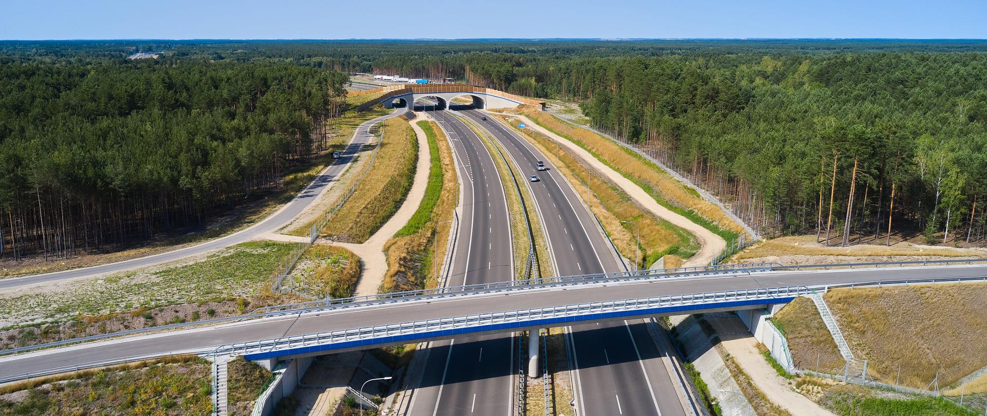 Zdjęcie lotnicze przedstawia nowoczesną drogę ekspresową przebiegającą przez rozległy kompleks leśny. Nad jezdniami znajdują się dwa wiadukty, a po obu stronach drogi widoczne są drogi serwisowe i skarpy. Na trasie porusza się kilka samochodów. Całość otaczają gęste zielone lasy, a nad horyzontem rozciąga się bezchmurne, błękitne niebo.
