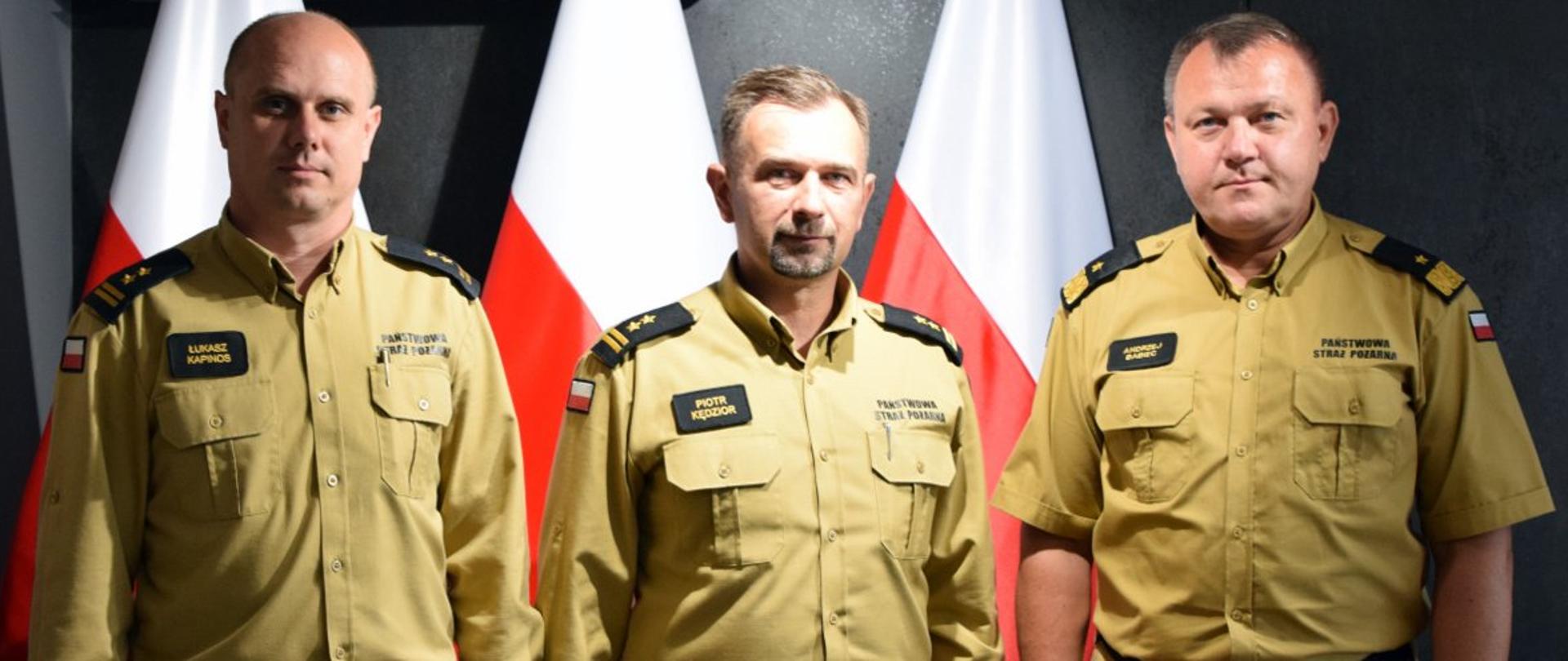 Zdjęcie zrobione wewnątrz pomieszczenia. Na zdjęciu trzech oficerów Państwowej Straży Pożarnej ubranych w mundury koloru piaskowego. Za nimi stoją biało-czerwone flagi.