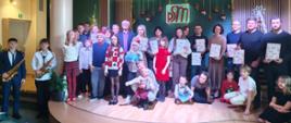 Na zdjęciu widać wszystkich uczniów biorących udział w koncercie którzy trzymają dyplomy w rękach i instrumenty muzyczne.
