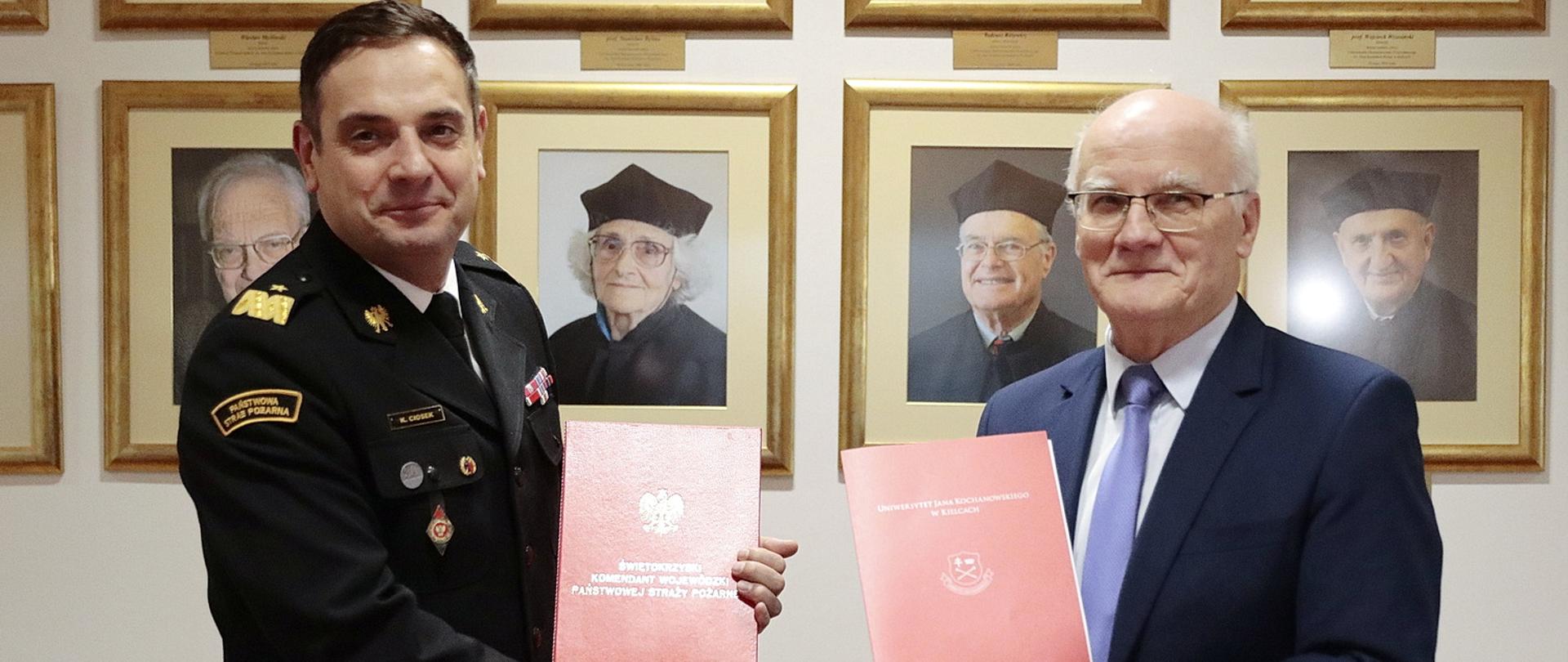 Na zdjęciu uścisk dłoni i wymiana teczek pomiędzy strażakiem w mundurze granatowym ze złotymi dystynkcjami generalskimi oraz rektorem UJK w garniturze. W tle zdjęcia z wizerunkami poprzednich rektorów UJK.