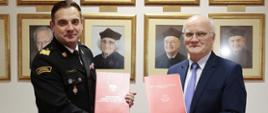 Na zdjęciu uścisk dłoni i wymiana teczek pomiędzy strażakiem w mundurze granatowym ze złotymi dystynkcjami generalskimi oraz rektorem UJK w garniturze. W tle zdjęcia z wizerunkami poprzednich rektorów UJK.