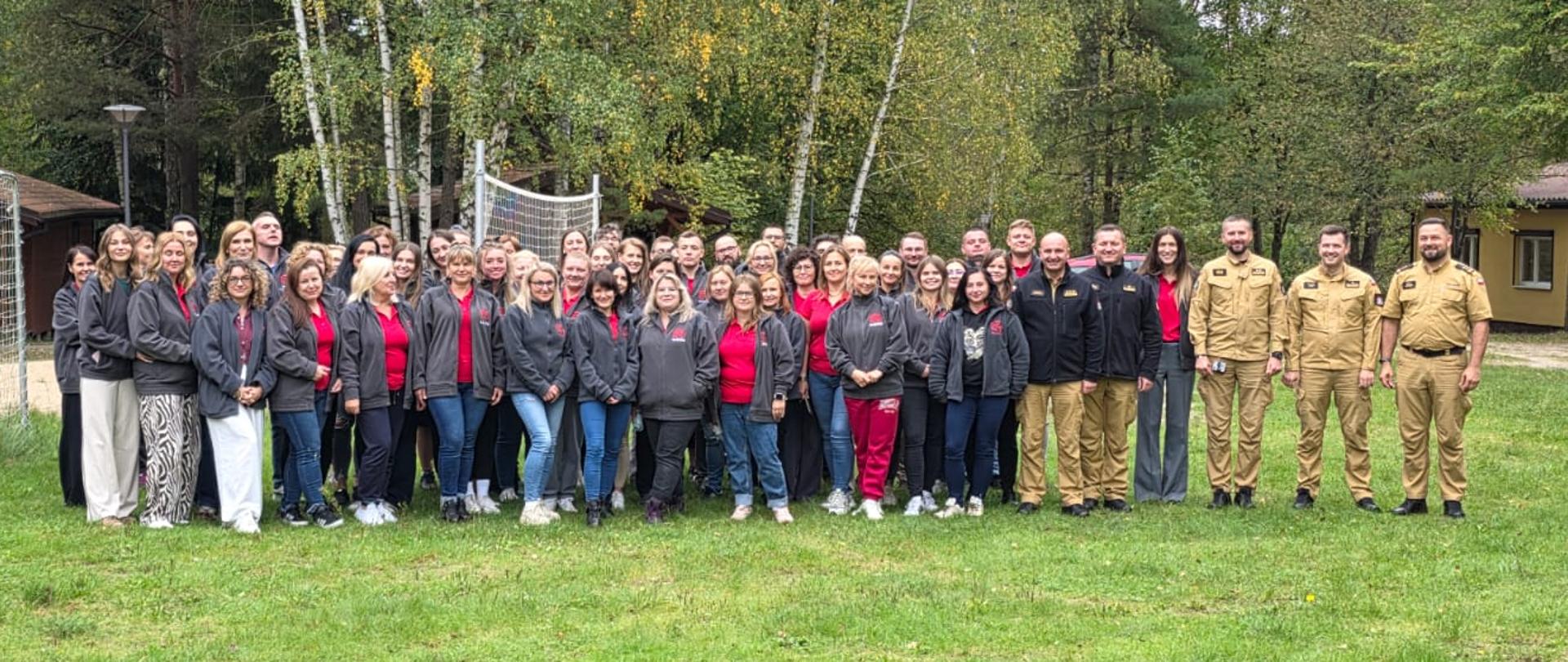 Na zdjęciu uczestnicy narady - kilku funkcjonariuszy PSP w piaskowych mundurach służbowych i pracownicy w czerwonych koszulkach i szarych polarach z napisem "112". Stoją na boisku sportowym, w tle fragment budynku i drzewa.