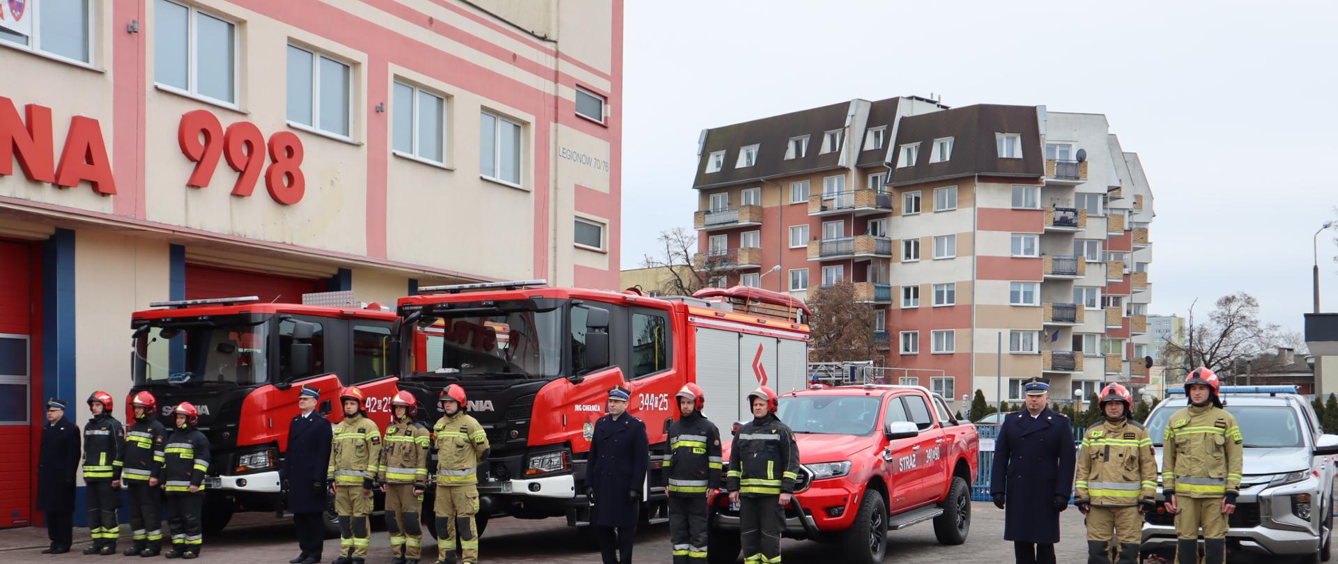 Strażacy na tle nowo przekazanych aut pożarniczych. Dwa ciężkie samochody ratowniczo-gaśnicze na podwoziu SCANIA z napędem 4x2 i dwa lekkie samochody ratowniczo-rozpoznawcze, jeden to: Ford Ranger o napędzie 4×4 i drugi to Mitsubishi L200 o napędzie 4x4. 