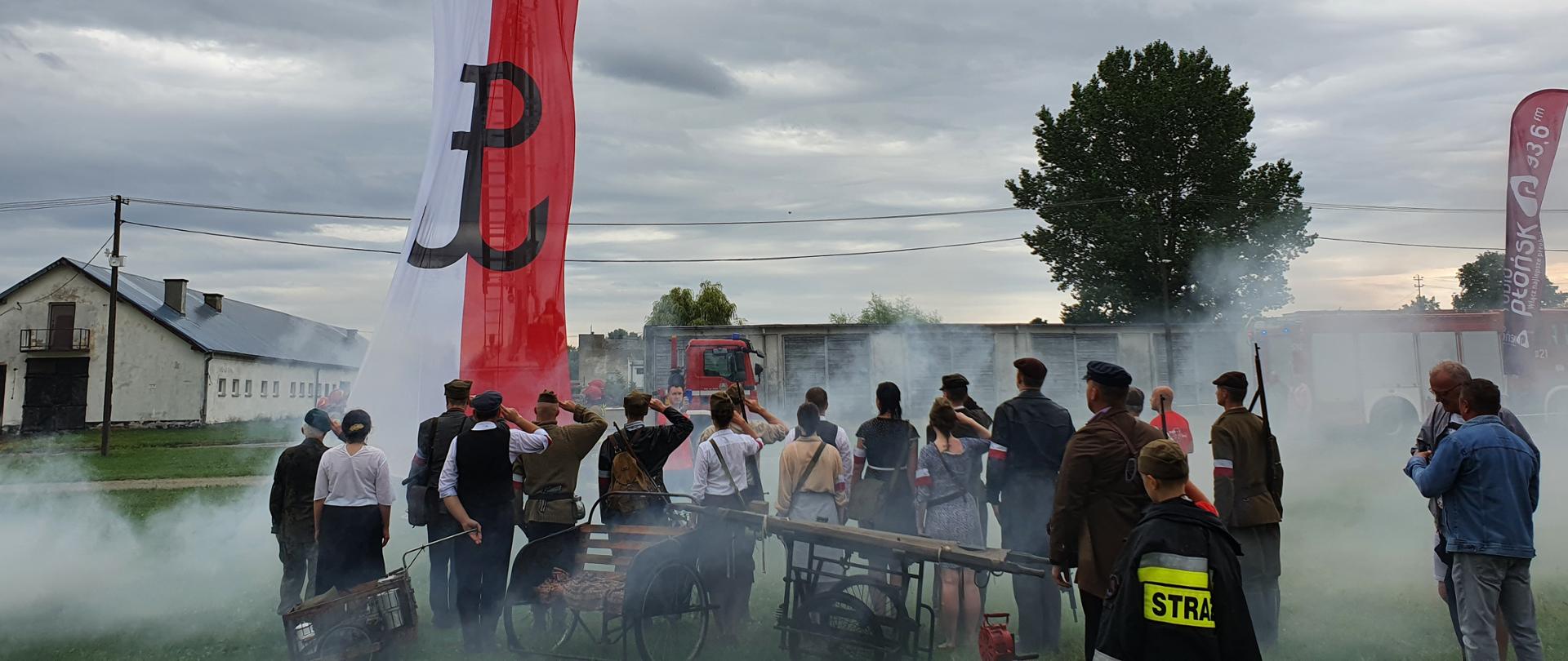 Flaga biało - czerwona ze znakiem Powstania Warszawskiego. Poniżej flagi grupa osób salutuje, skierowana w kierunku flagi.