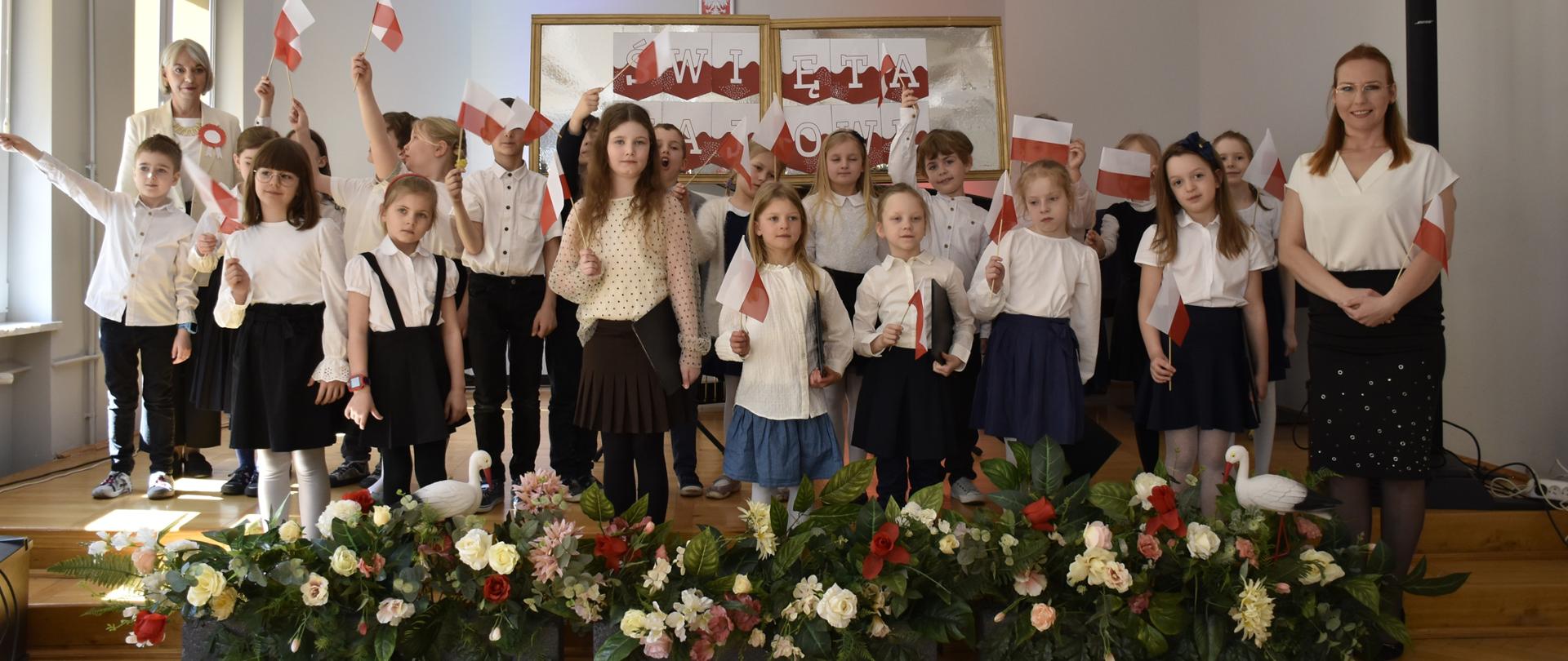 na pierwszym planie dzieci z klasy I z flagami białoczerwonymi w tle biało czerwony napis święta majowe