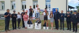Zdjęcie przedstawia stojących na podium biegaczy trzymających puchary. Po prawej i lewej stronie organizatorzy zawodów i zaproszeni goście.