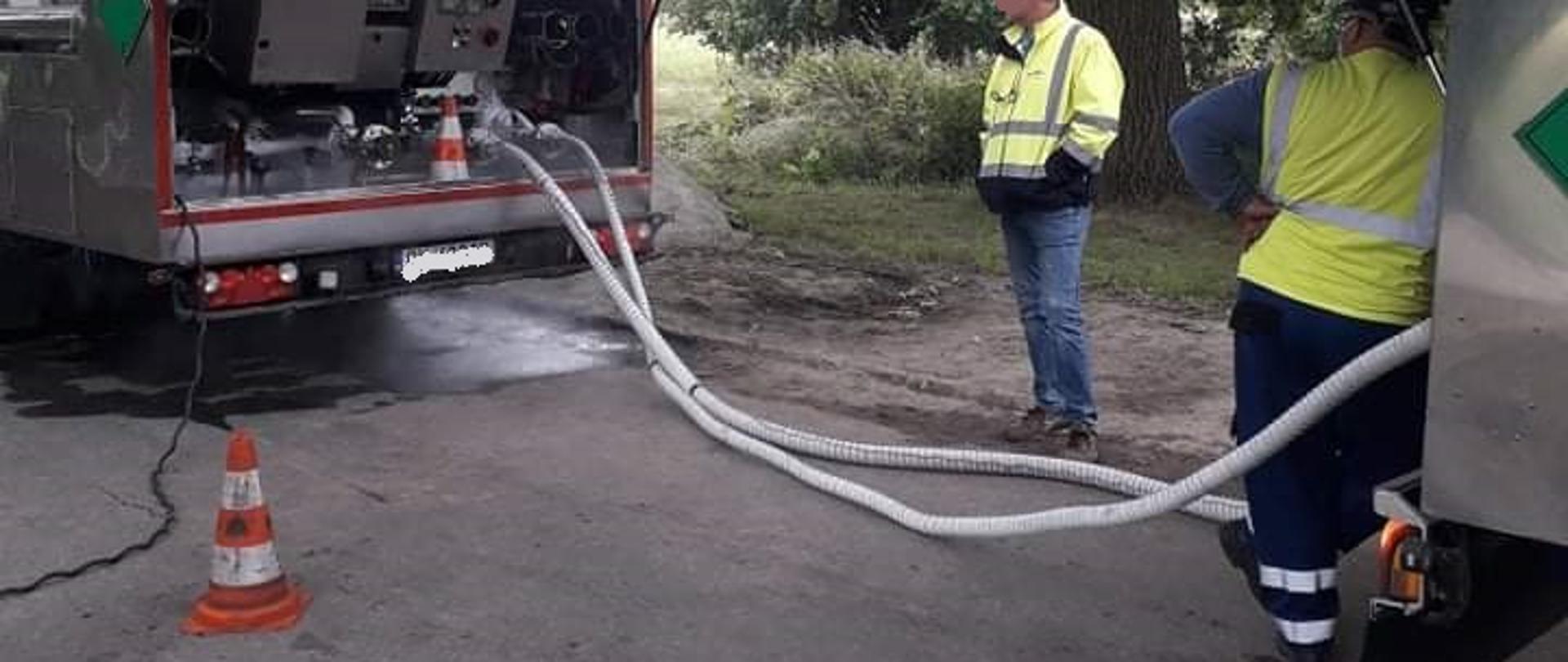 Na środku zdjęcia widoczni są dwaj mężczyźni, którzy stoją na asfaltowej drodze ubrani w żółte bluzy i granatowe spodnie oraz białe kaski na głowach. Kontrolują przeładunek dwutlenku węgla. Po prawej stronie zdjęcia widoczna jest tylna skrzynia od cysterny koloru srebrnego o którą oparty jest jeden z mężczyzn. Po lewej stronie zdjęcia w lewym dolnym rogu stoi na asfalcie stożek ostrzegawczy koloru pomarańczowo – białego. W dalszym planie widoczna jest cysterna koloru białego , na której widoczne są zielono niebieskie napisy . Z tyłu cysterna posiada srebrną skrzynię , z której poprowadzone są białe węże do przepompowania dwutlenku węgla. W oddali widać dwa drzewa drzewa.