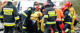 Na zdjęciu z odległości kilku metrów widać rozbity samochód osobowy marki Audi oraz strażaków, którzy wykonują dostęp do kierowcy tego samochodu - jest on zakleszczony. W oddali widać pojazdy ratownicze. Aura deszczowa.