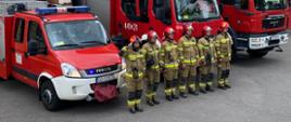Strażacy ustawieni w szeregu przed pojazdami z załączoną sygnalizacją