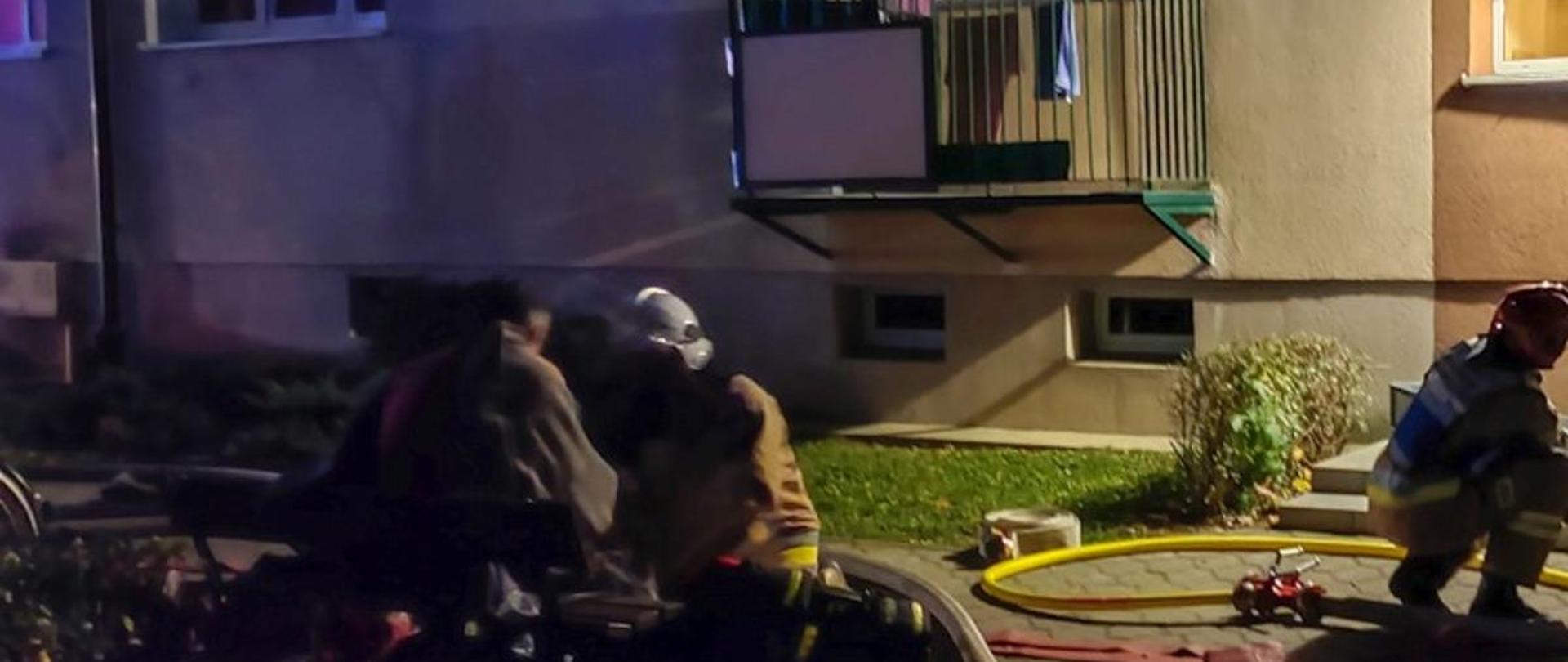 Na zdjęciu 3 strażaków przed klatką i na trawniku, rozwinięte linie gaśnicze oraz wentylator oddymiający 