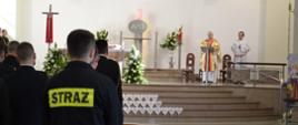 Strażak w ubraniu koszarowym po lewj stronie kadru stoi tyłem, na plecach ubrania napis STRAŻ, po prawej stronie kadru ksiądz -celebrujący mszę św. W tle chody do ołtarza
