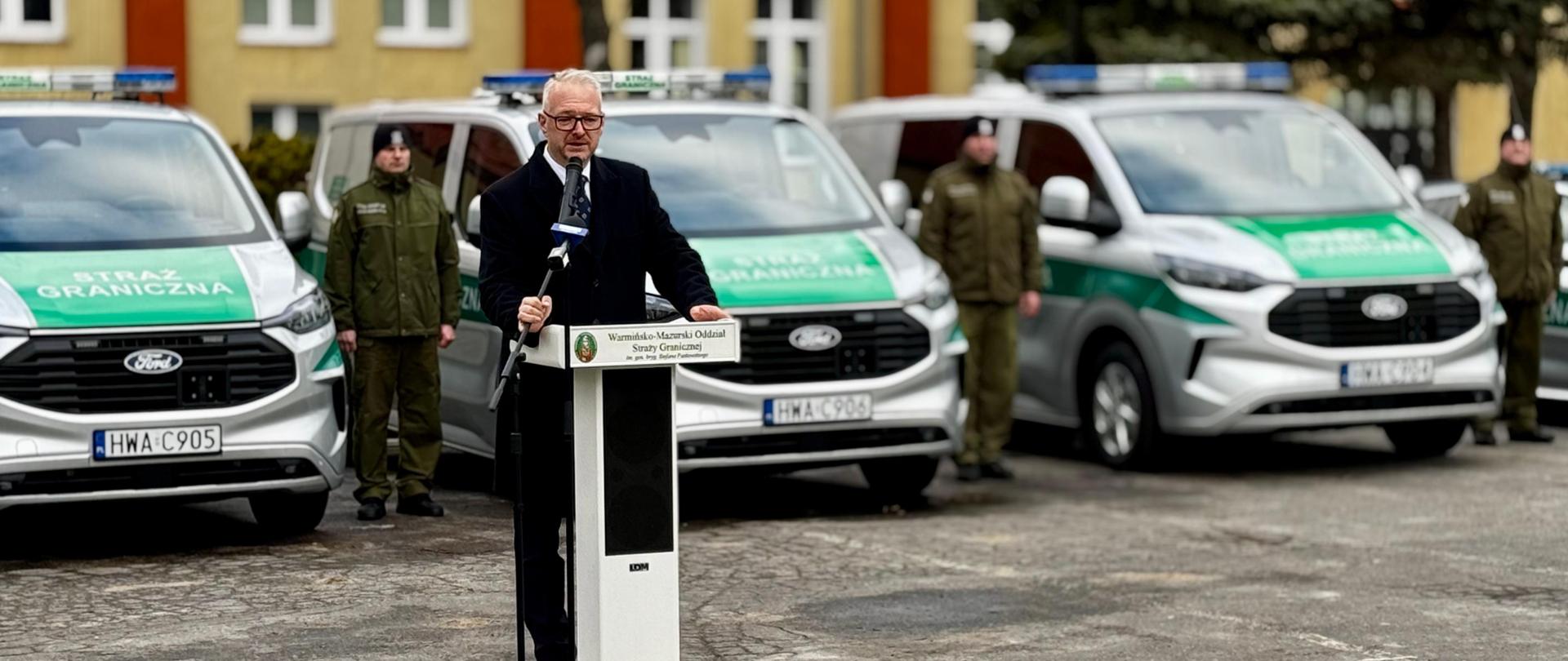 Wojewoda stoi przy mównicy na tle aut Straży Granicznej 