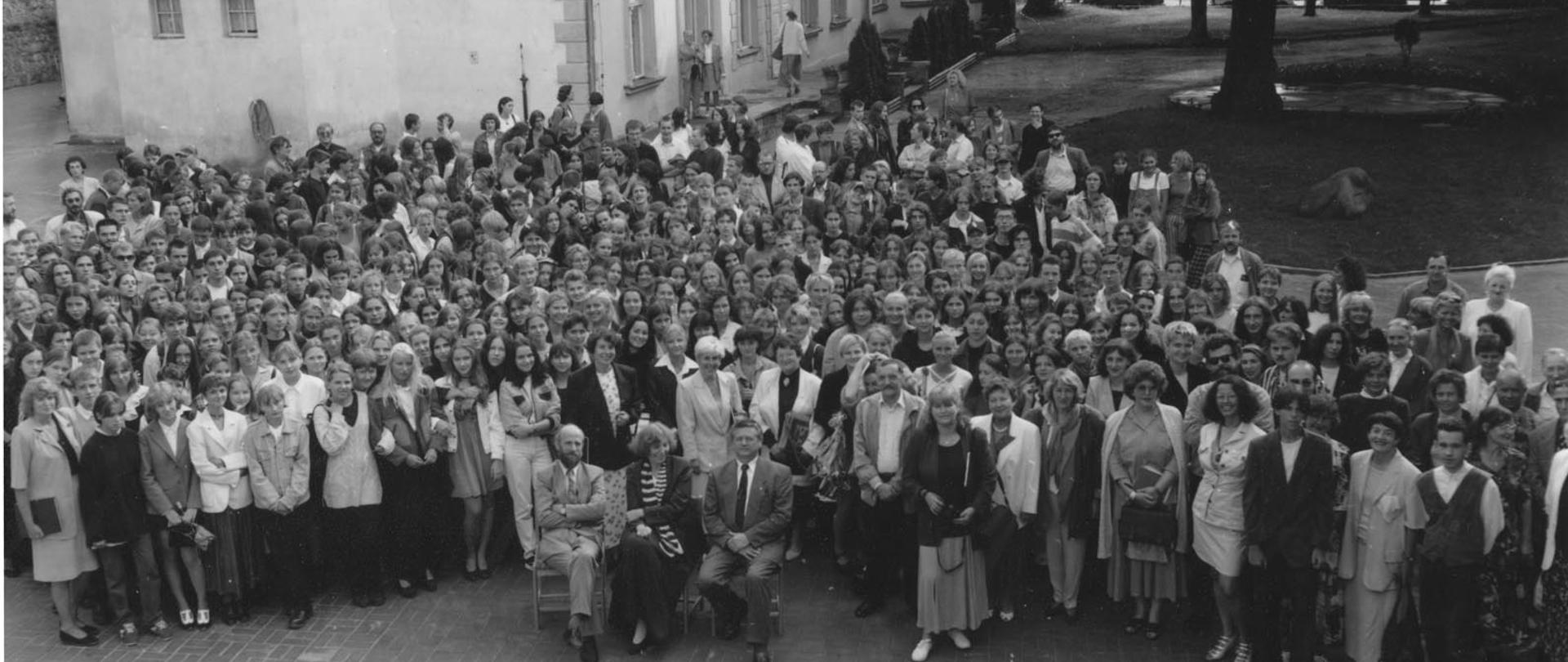 Czarno-biała fotografia wykonana z wysokości 1. piętra przedstawia grupę ok. 300 osób na rozpoczęciu roku szkolnego. W tle stary budynek szkoły i fragment parku przed budynkiem.