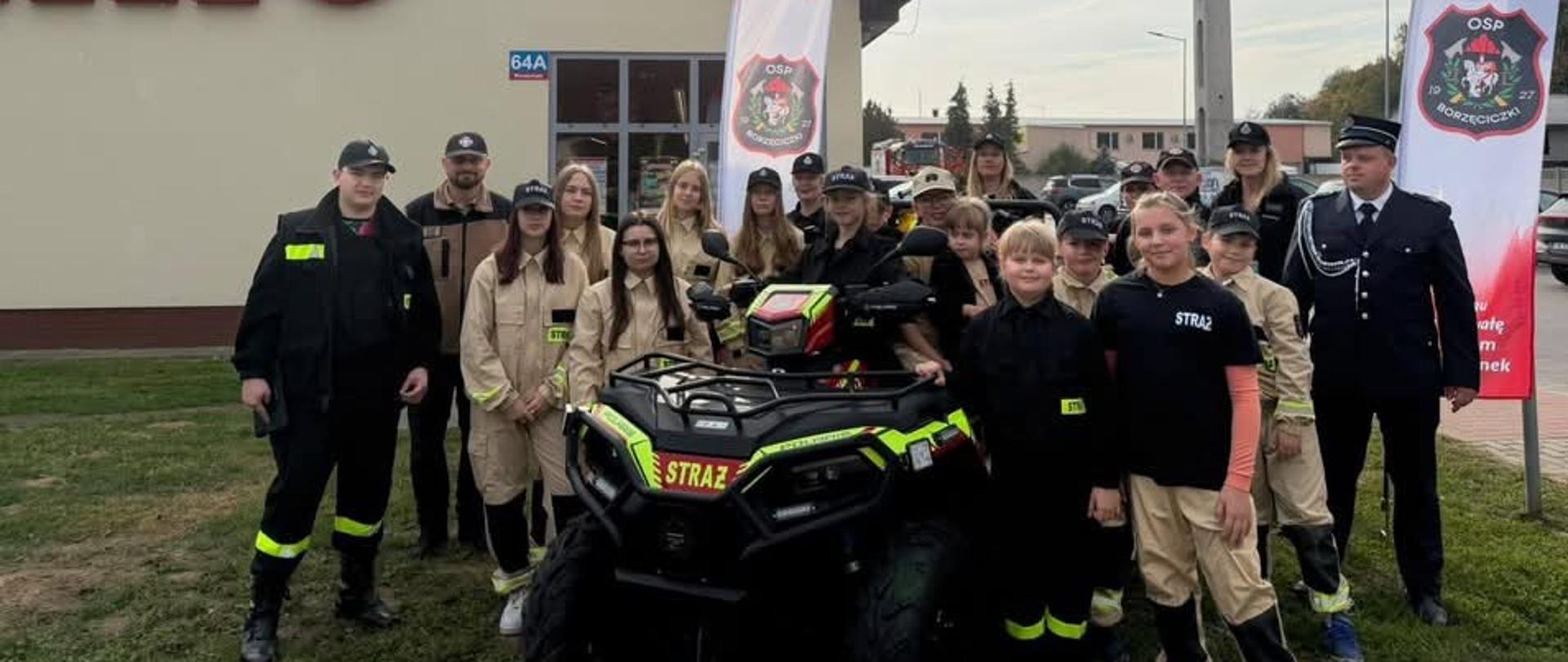 Zdjęcie grupowe członków jednostki OSP w umundurowaniu na tle miejsca publicznego. Na pierwszym planie widać sprzęt w postaci quada. 