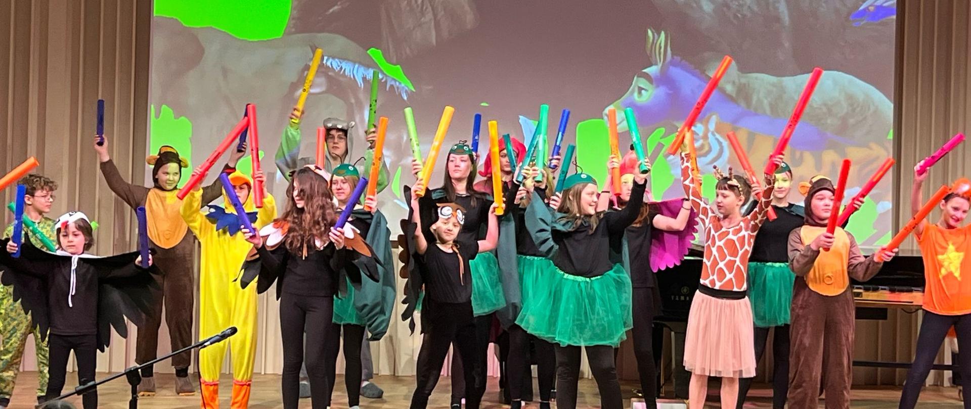 Na zdjęciu uczniowie przebrani za zwierzęta, którzy grają na instrumentach. Zdjęcie zostało wykonane podczas koncertu rekrutacyjnego. W tle na rzutniku grafika przedstawiająca zwierzęta. Podłoga drewniana, a tło obrazka w odcieniach beżu.