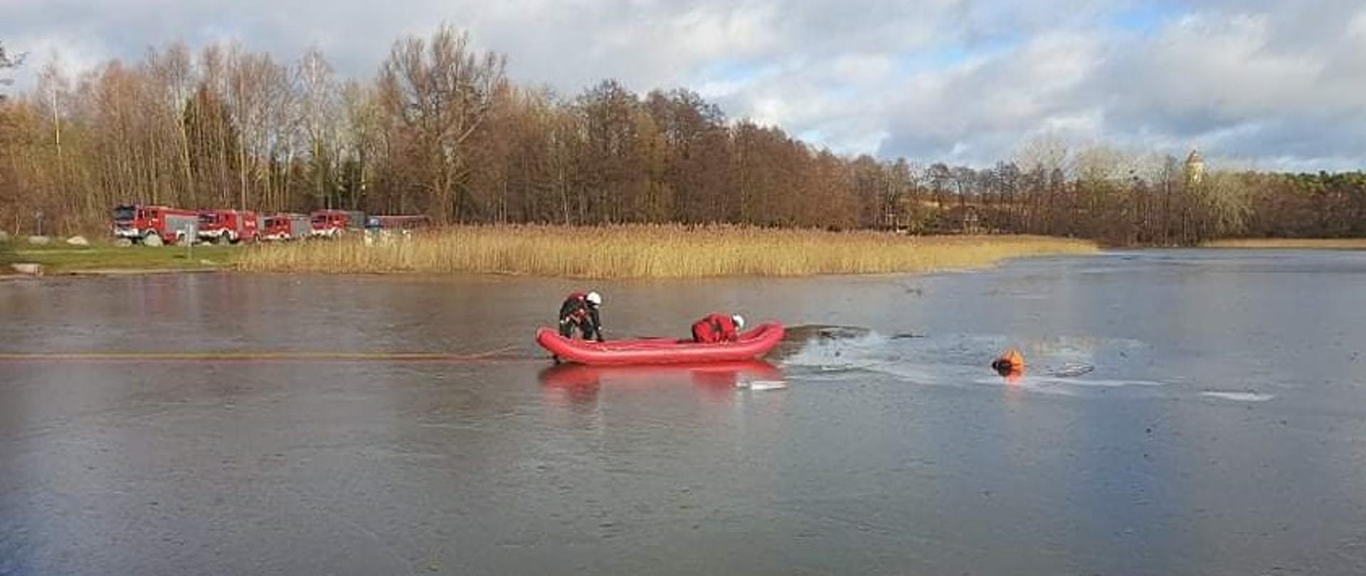 Na zamarzniętym akwenie wodnym strażacy ćwiczą ewakuację osoby poszkodowanje z przerebla. W oddali za nimi widać samochody ratowniczo gaśnicze i trzcinę.