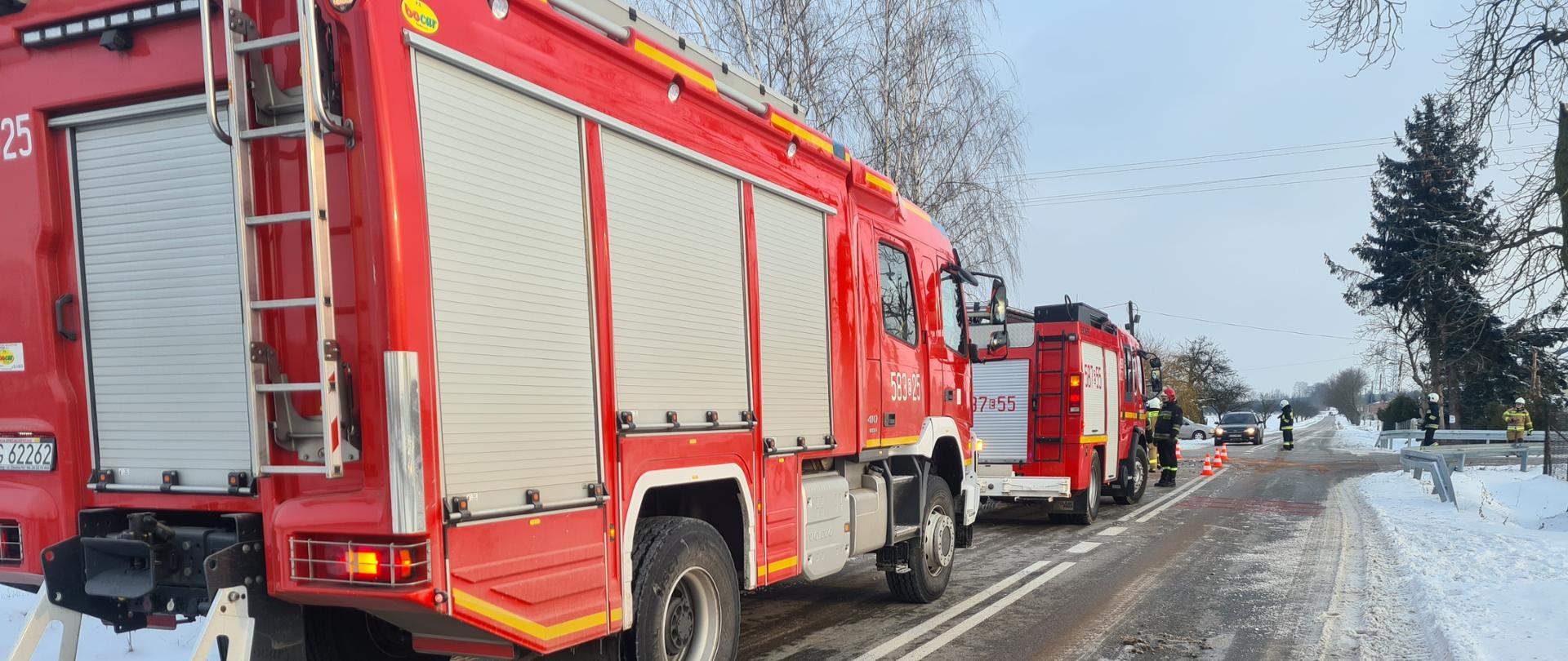 Na zdjęciu widać dwa samochody ratowniczo gaśnicze zabezpieczające miejsce kolizji. W oddali widać 2 strażaków kierujących ruchem.