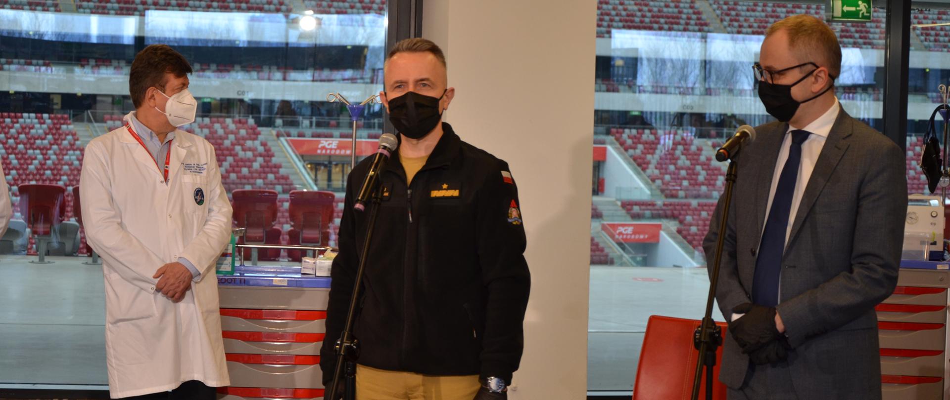 Na zdjęciu widzimy dwóch mężczyzn w średnim wieku stojących przy mikrofonach. Mężczyzna po prawej ubrany w ciemno-szary garnitur, granatowy krawat, na rękach czarne rękawiczki, na twarzy czarne maseczka i okulary w czarnej ramce. Środek kadru to drugi mężczyzna strażak, ubrany w mundur, spodnie koloru piaskowego, bluza koloru czarnego. Mężczyzna na rękach czarne rękawiczki, zegarek na lewej ręce, czarna maseczka na twarzy. Lewa strona kadru to mężczyzna w białym kitlu i czarnych spodniach na twarzy biała maseczka. Mężczyźni stoją w pomieszczeniu białe ściany, brązowa podłoga. Dalej w kadrze widzimy wielkie okna od podłogi aż po sufit, za oknami widzimy trybuny stadionu i biało-czerwone krzesełka. 