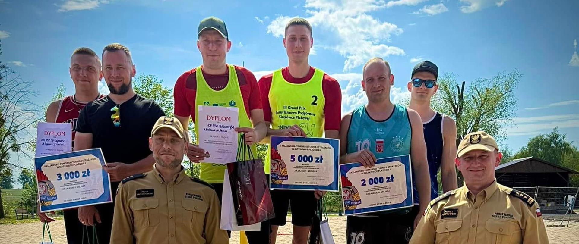 Mistrzostwa Województwa Kujawsko-Pomorskiego Strażaków PSP w Plażowej Piłce Siatkowej. Zdjęcie przedstawia strażaków PSP podczas wręczenia nagród zawodnikom. W tle strażacy PSP na podium.