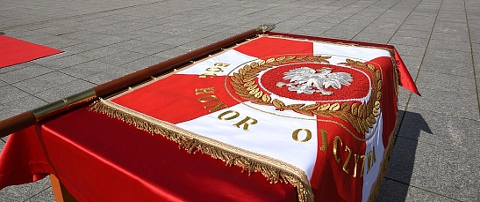 Sztandar dla wągrowieckich strażaków leżący na stole, w tle grupy ludzi stojących na placu