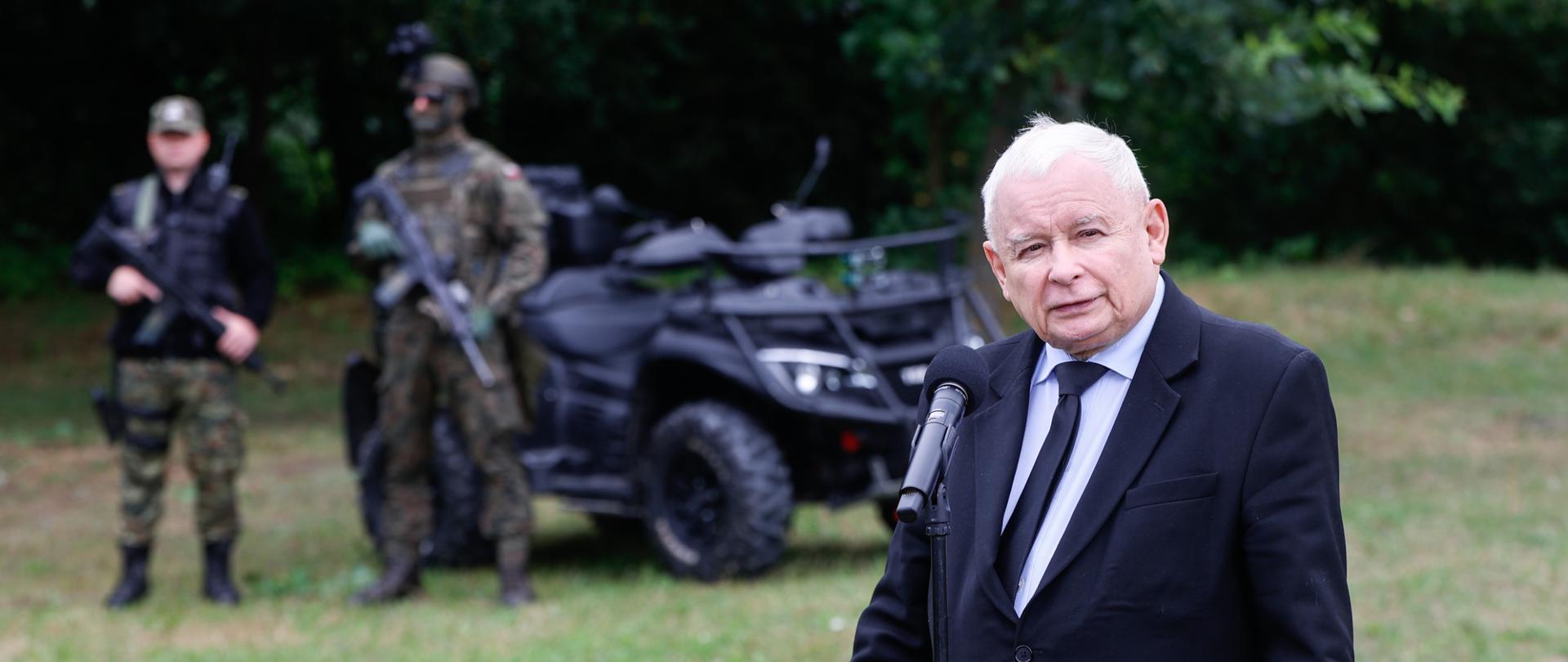 Wicepremier stoi przy mikrofonie. W tle samochód i dwóch strażników Straży Granicznej.