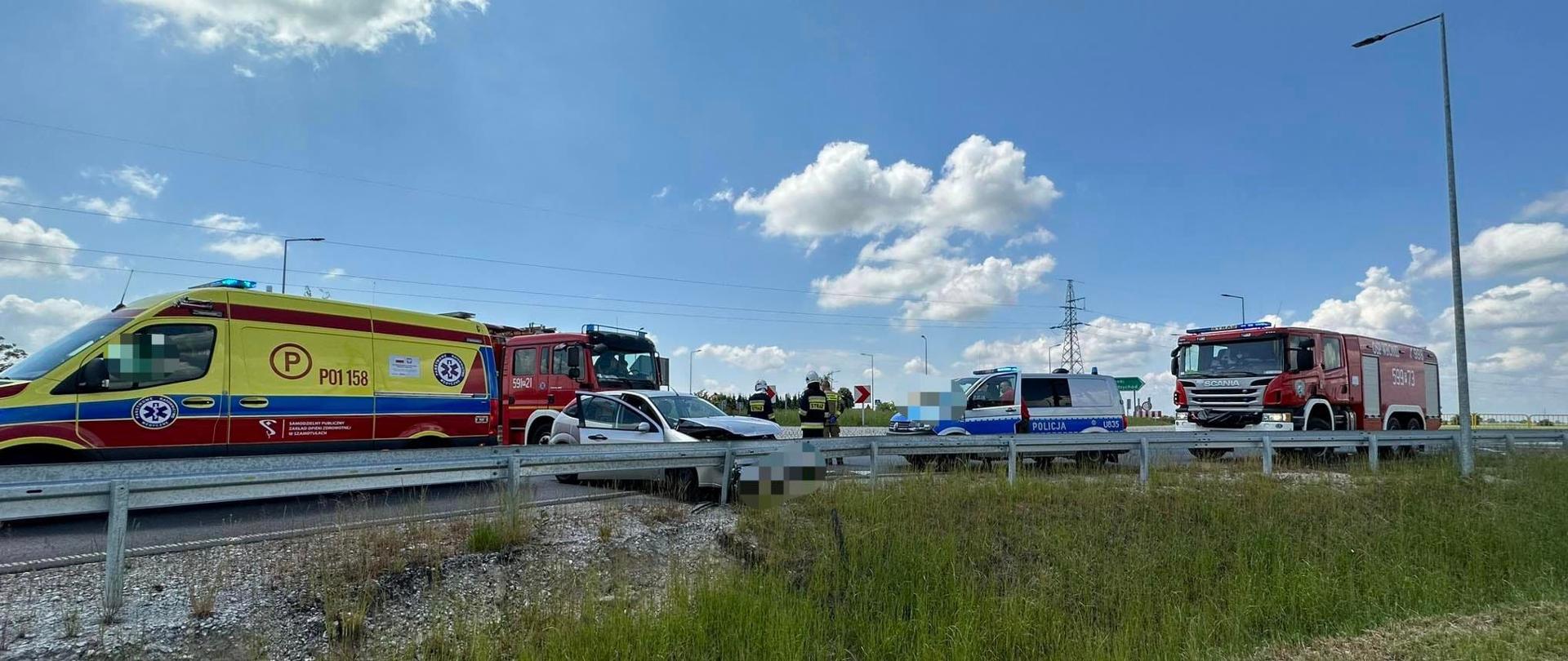 Po lewej widać karetkę pogotowia, po prawej uszkodzony pojazd przy barierkach, za nim policja i straż pożarna