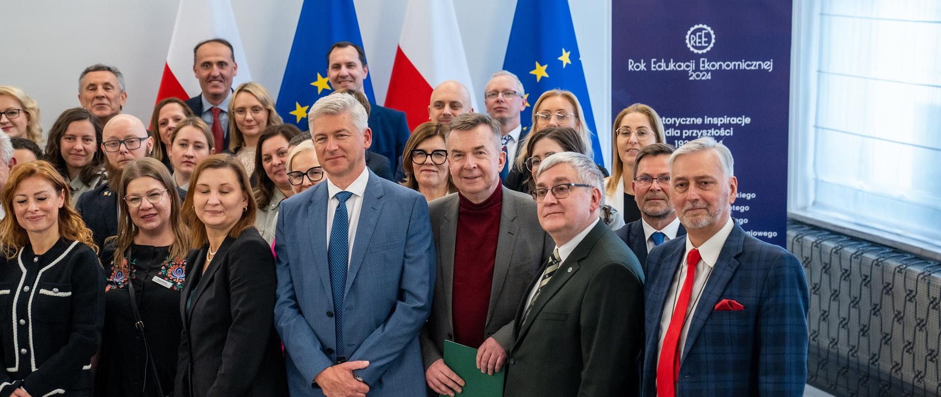 Zdjęcie zbiorowe, na sali stoi duża grupa osób, za nimi pod ścianą flagi Polski i UE.
