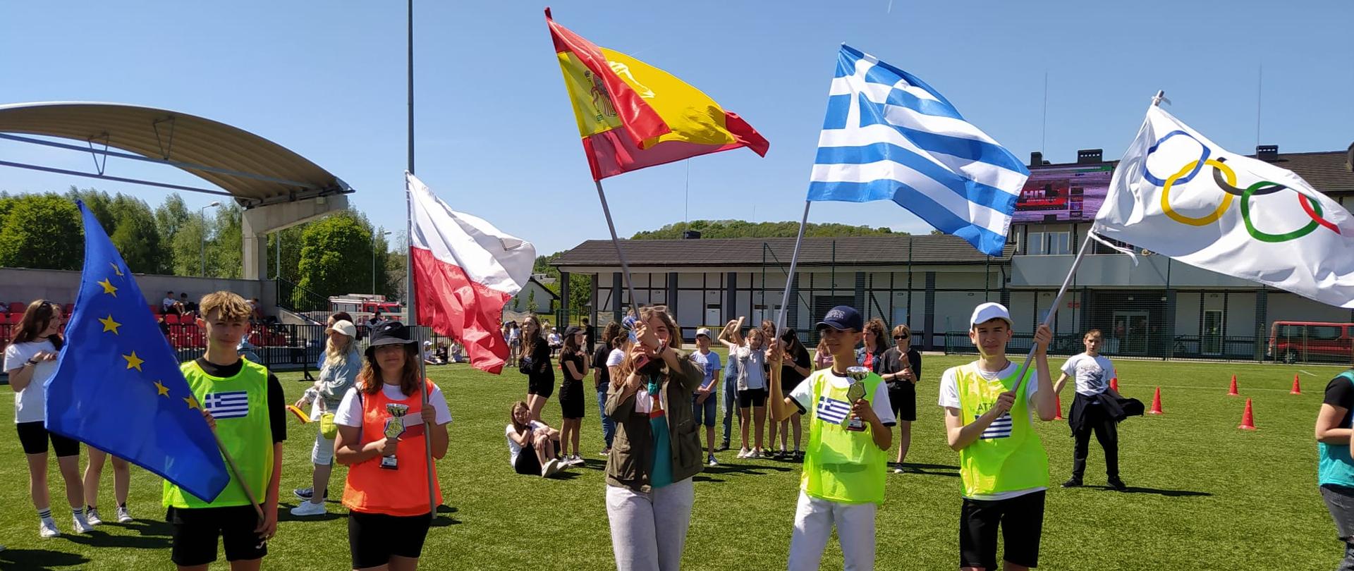Na zdjęciu dzieci z flagami Hiszpanii, Grecji, Polski, Unii oraz olimpijską wraz z pucharami na tle stadionu piłkarskiego