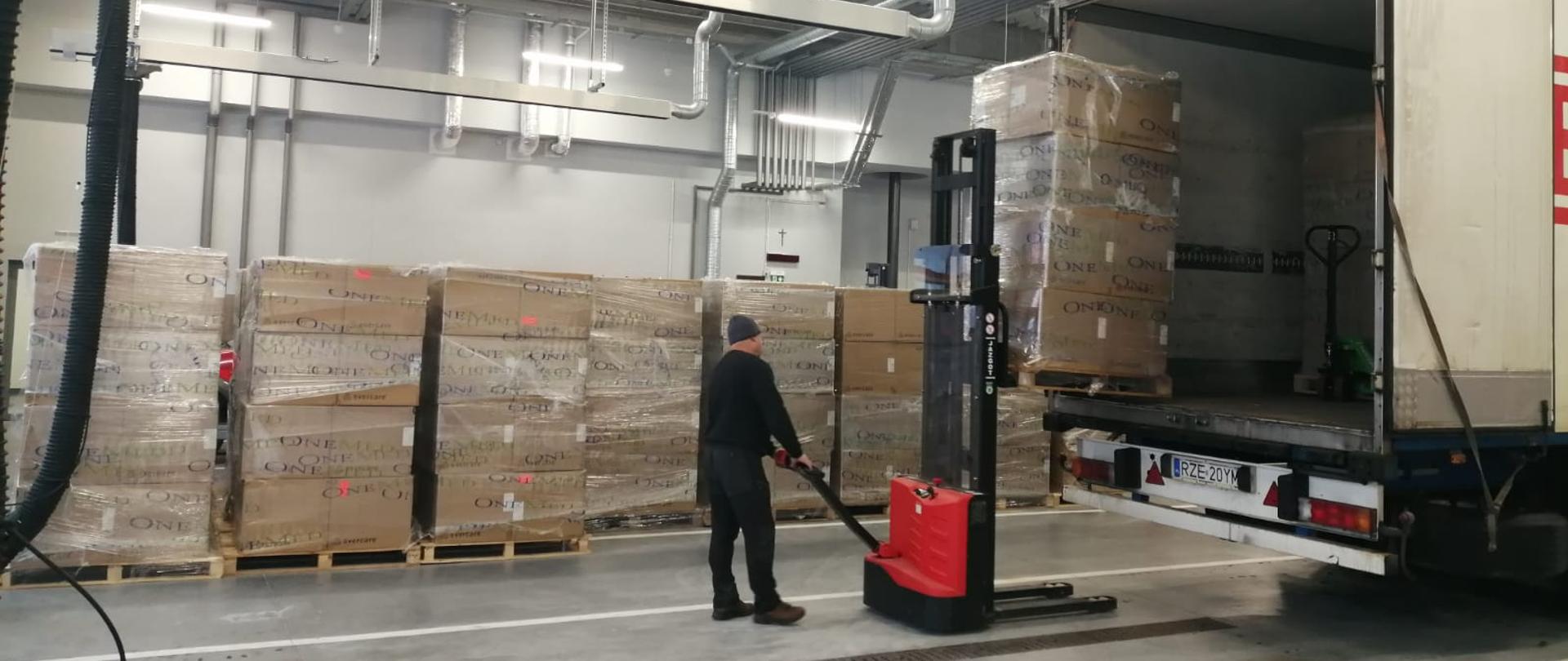 Zdjęcie przedstawia wnętrze garażu Jednostki Ratowniczo-Gaśniczej Nr 3 w Rzeszowie, w którym odbywa się rozładunek tira dostarczającego maseczki chirurgiczne oraz typu FFP2 przy użyciu wózka widłowego. Pod ścianą garażu widać rozładowane palety z rozładowanymi kartonami.