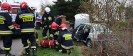 Widoczny rozbity samochód, ratownicy PSP i OSP, ratownicy medyczni oraz samochody służb ratowniczych.