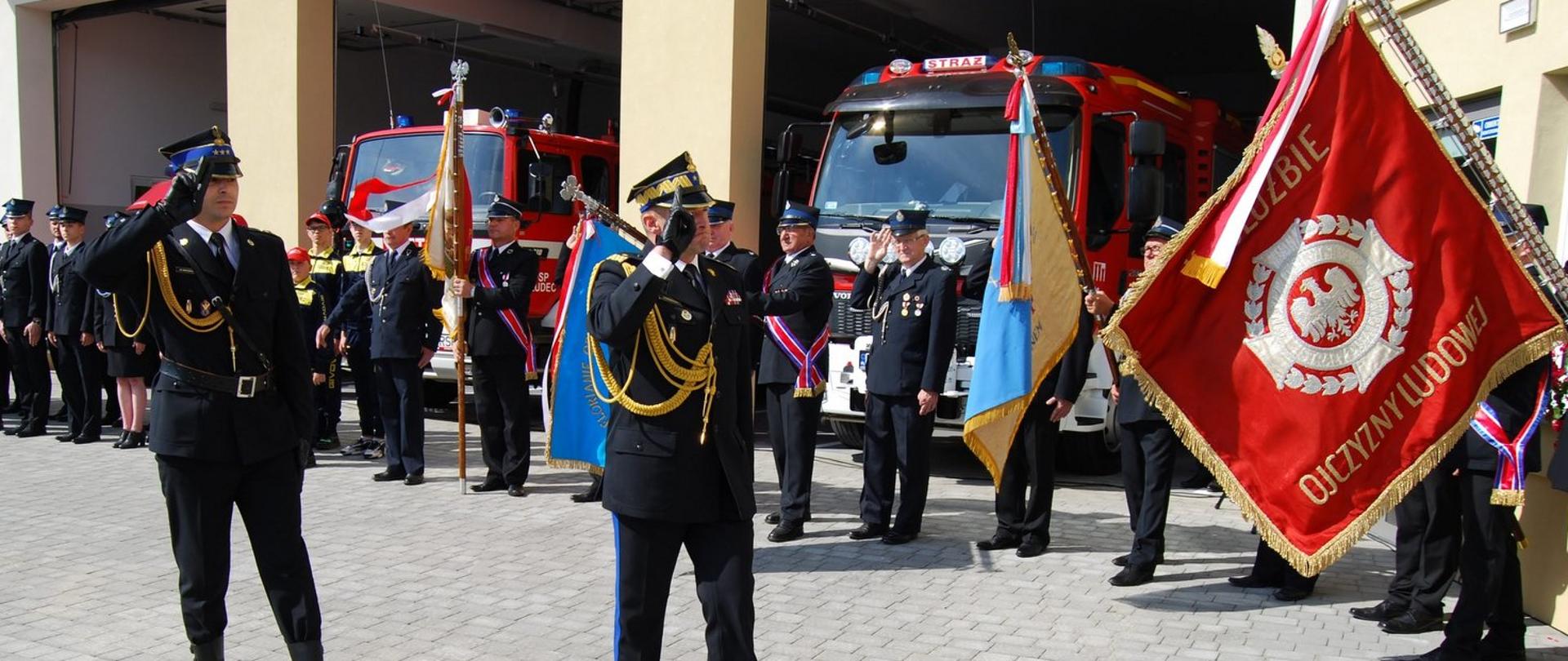 Zdjęcie zrobione przed remizą strażacką. Na pierwszym planie widać dwóch oficerów Państwowej Straży Pożarnej obydwaj salutują w stronę strona sztandaru, który jest widoczny po prawej stronie zdjęcia. Jeden z oficerów jest w stopniu generała. Za nimi widać ustawionych w dwuszeregu strażaków ochotników, za którymi stoją dwa samochody pożarnicze.