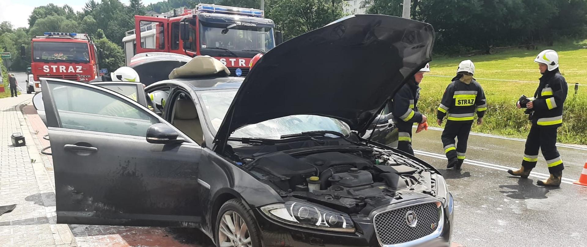 Samochód w którym powstał pożar na zatoczce autobusowej z otwartymi drzwiami oraz klapa silnika. Na drodze leżą zużyte gaśnice proszkowe. Obok samochodu strażacy.