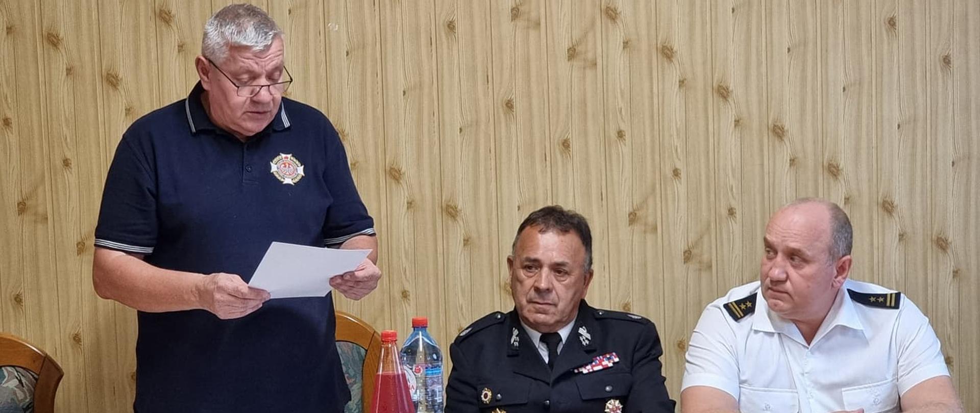 Na zdjęciu widać druhów z OSP oraz przedstawiciela Komendy Powiatowej Państwowej Straży Pożarnej w Ustrzykach Dolnych którzy siedzą przy stole a jeden z druhów przemawia czytając z kartki na stojąco. 