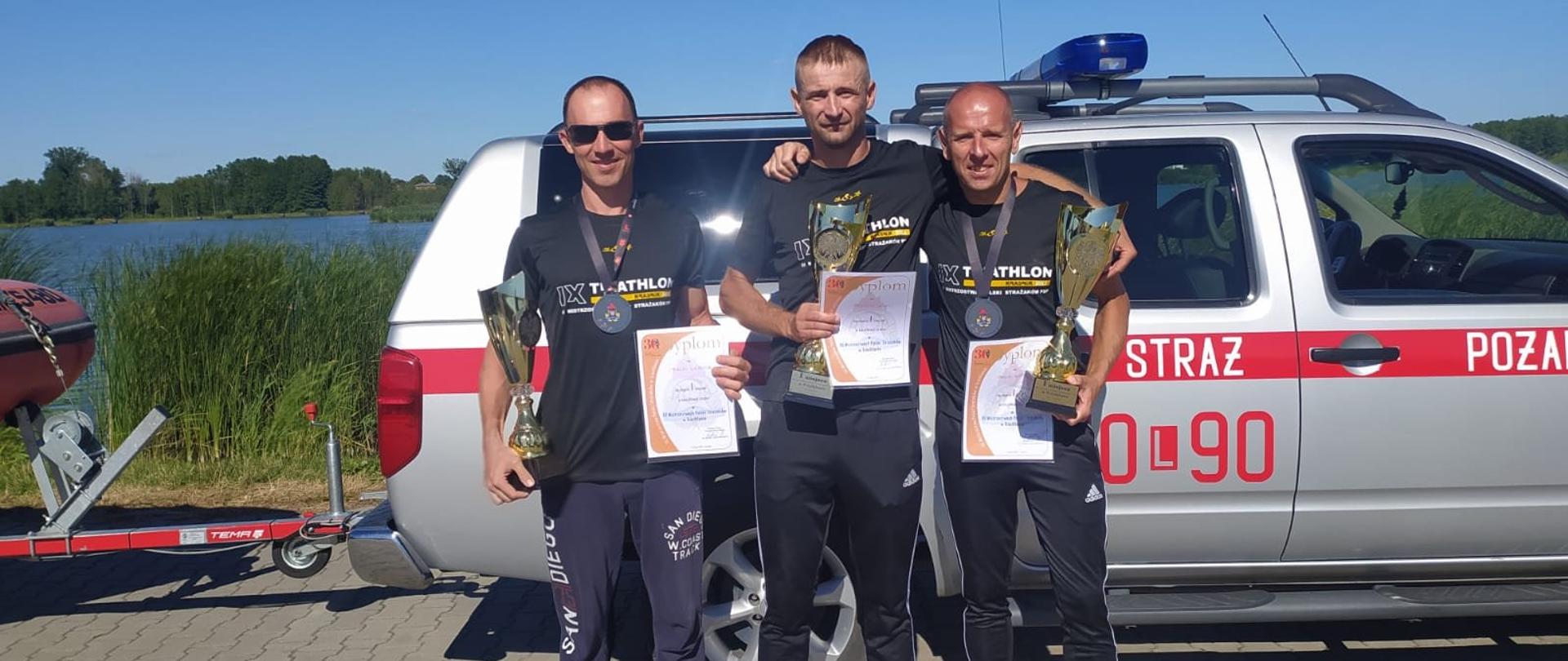 Trzech strażaków w strojach sportowych trzymających dyplomy i puchary w rękach