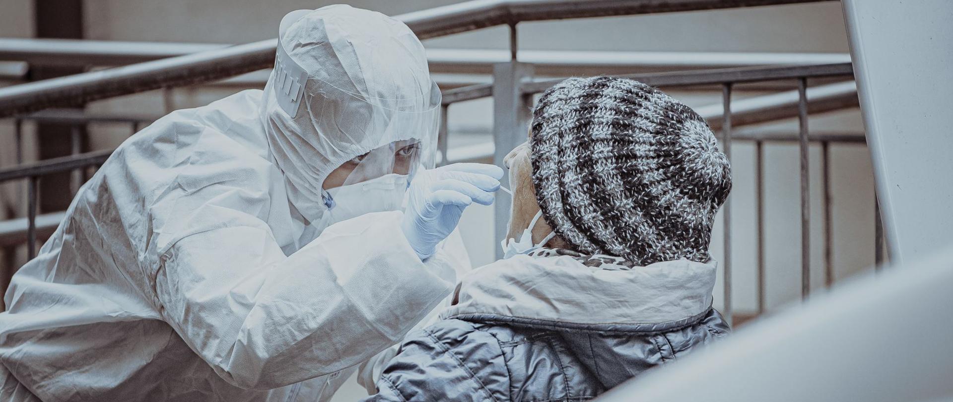 Na zdjęciu widać lekarza w białym kombinezonie, który pobiera wymaz (badanie) od starszej kobiety