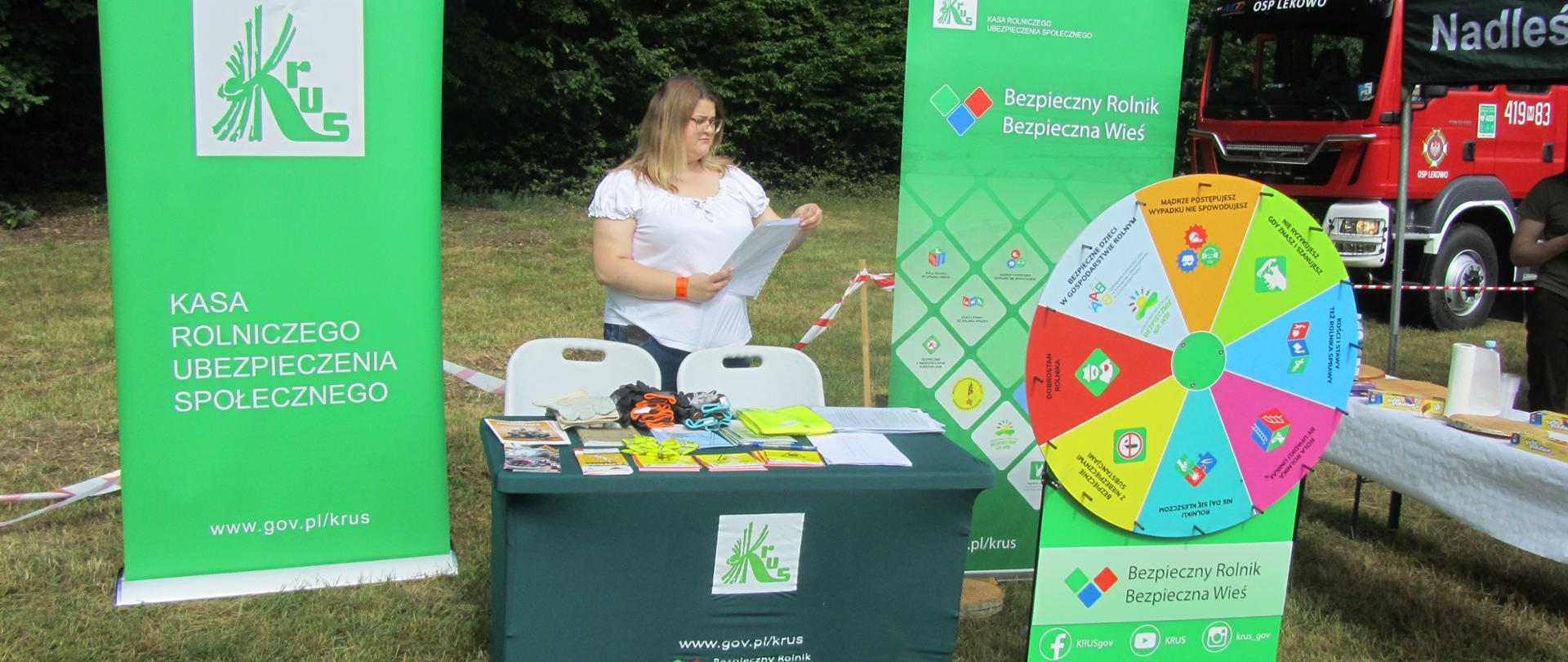 Stoisko informacyjno-prewencyjne przygotowane przez PT KRUS w Ciechanowie. Stolik przyozdobiony zielonym obrusem z logiem Kasy współgra kolorystycznie z banerami oraz kołem prewencyjnym. Kobieta stojąca przy stoisku w skupieniu przygotowuje dokumenty dla uczestników gry prewencyjnej.