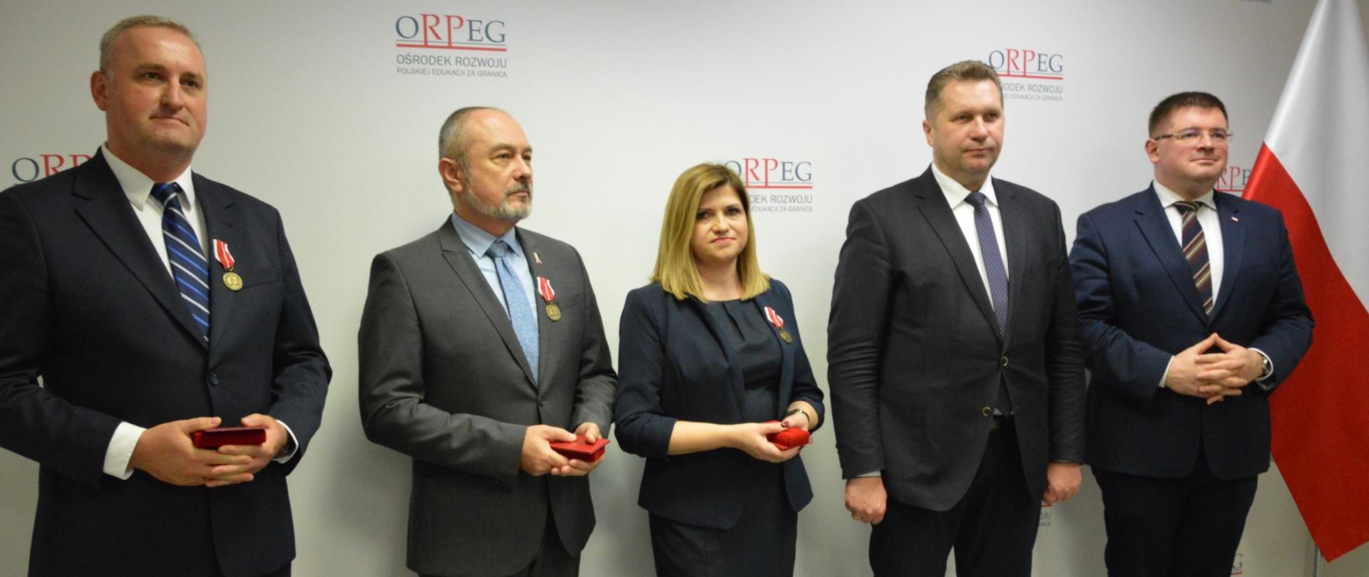 Uczestnicy uroczystości stoją pod ścianą, obok nich polska flaga.