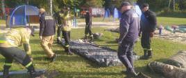 Biwak klas mundurowych. Zdjęcie przedstawia teren biwaku, na którym odbywa się rozbijanie namiotów. Czynności te wykonują strażacy oraz funkcjonariusz Służby Więziennej . W tle urządzenia placu zabaw dla dzieci oraz wóz strażacki.
