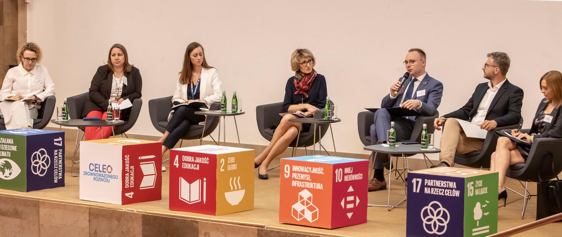 VI edycja Krajowego Forum Interesariuszy Agendy 2030. Na zdjęciu widać uczestników Forum, którzy siedzą na scenie. Przed nimi kolorowe, kwadratowe pufy.
