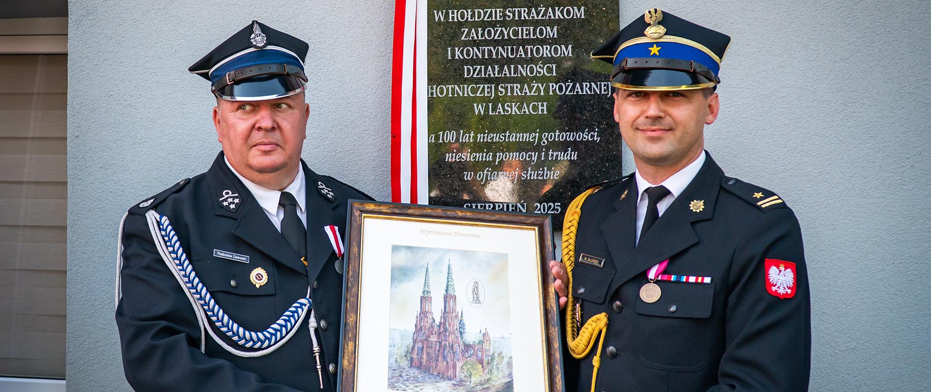 Dwóch mężczyzn w mundurach strażackich stoi obok tablicy pamiątkowej z napisem upamiętniającym 100-lecie OSP Laski. Jeden z nich trzyma w rękach oprawiony obraz z katedrą i napisem „Wyróżnienie Honorowe”. Mężczyźni mają poważny, ale dumny wyraz twarzy. Tło stanowi ściana budynku, tablica i biało-czerwona wstążka.