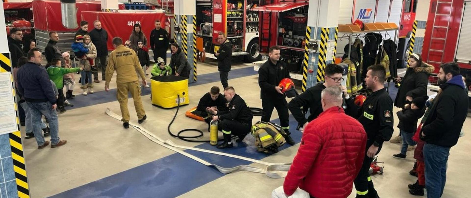 Na zdjęciu w ramach akcji "Otwartej Strażnicy" z okazji 32 Finału Wielkiej Orkiestry Świątecznej Pomocy osoby dorosłe i dzieci zwiedzają Jednostkę Ratowniczo-Gaśniczą. Widoczni na zdjęciu na garażu jednostki strażacy prezentują sprzęt pożarniczy przybyłym na akcję osobom. 