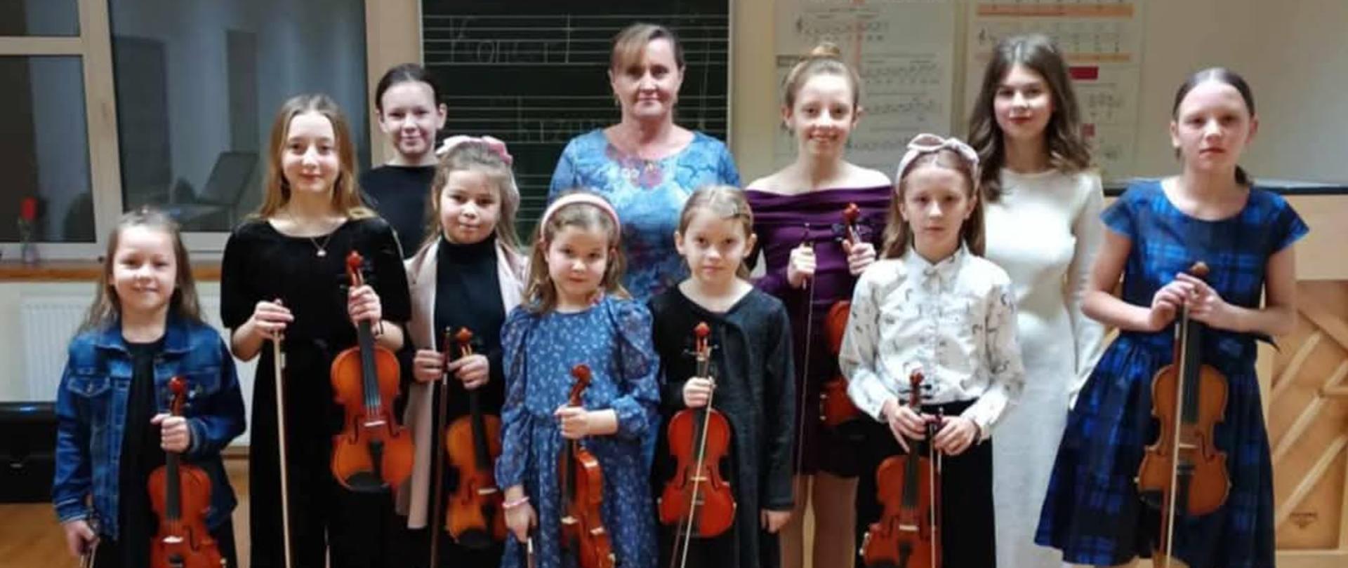 Zdjęcie zbiorowe na którym są same dziewczyny. Wszystkie trzymają skrzypce, są odświętnie ubrane. Wśród nich jest ich nauczycielka. Wszyscy uśmiechają się.