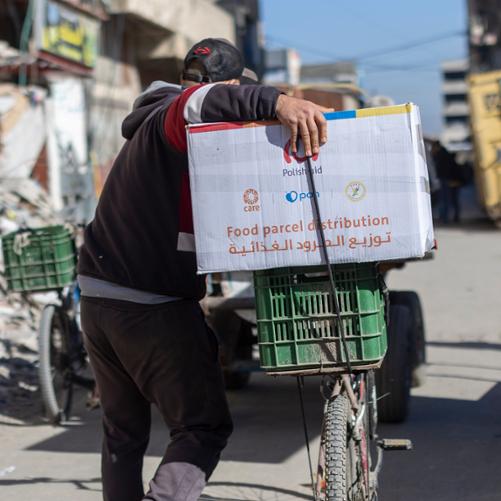 Na ulicy pełnej zniszczonych budynków mężczyzna prowadzi rower, na którym ustawiona jest paczka z pomocą żywnościową