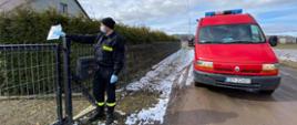 W lewej części zdjęcia strażak w ubraniu koszarowym czarnym, na głowie czapka zimowa, na twarzy maseczka. Strażak trzyma prawej wyprostowanej ręce ulotkę i patrzy w lewą stronę stojąc przy płocie posesji. W prawej części zdjęcia przód pojazdu pożarniczego marki Renault należący do OSP Gościeradz. 