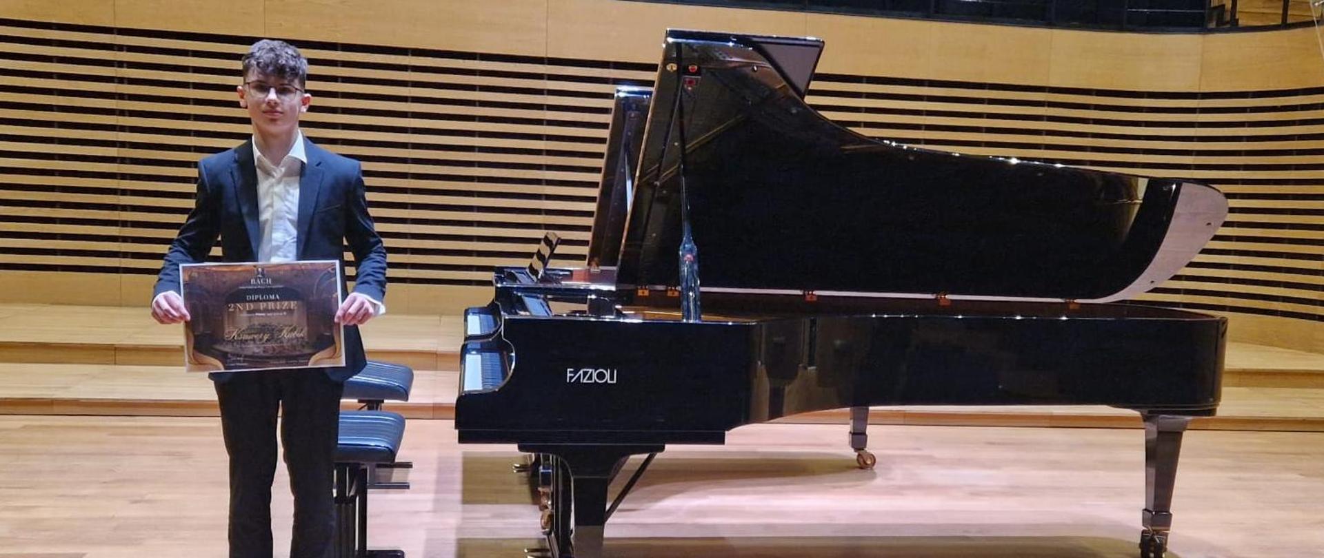 Ksawery Kubik stojący obok fortepianu z dyplomem z konkursu