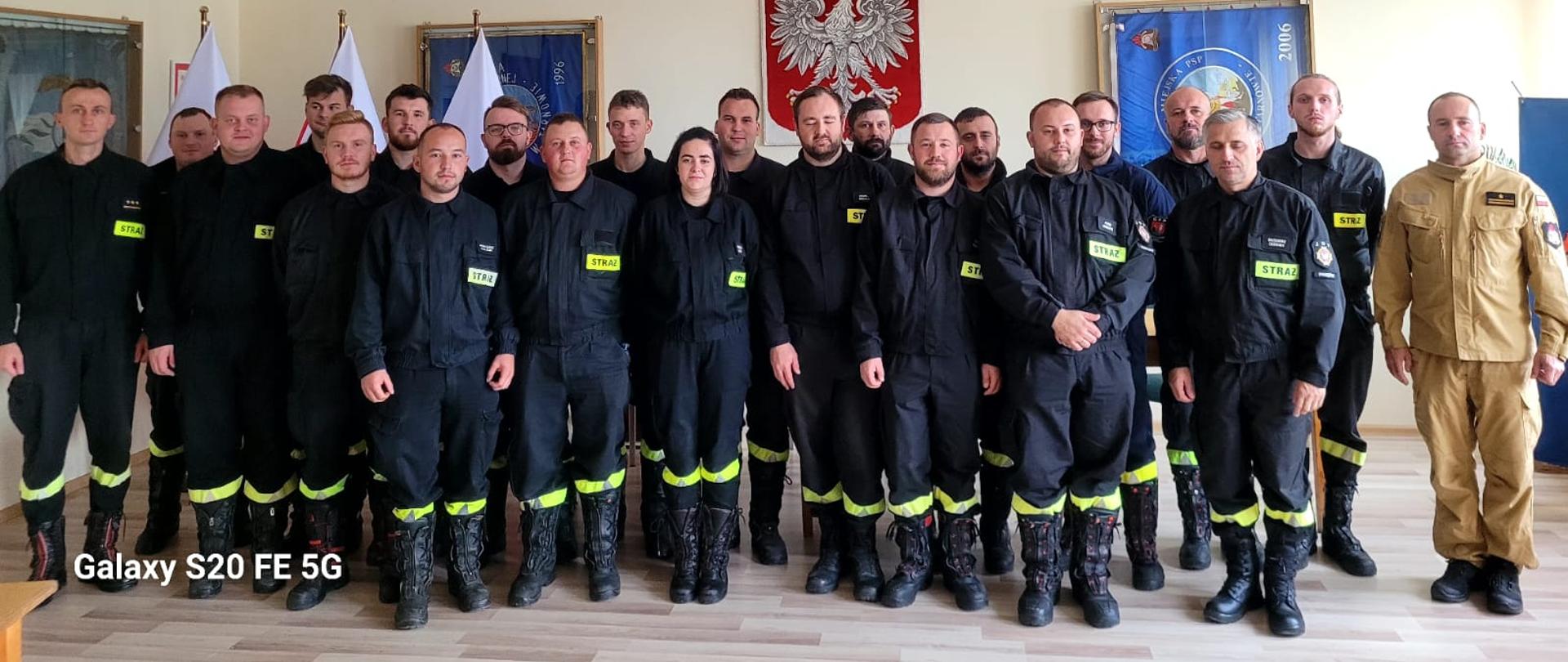 Na zdjęciu są widoczni stojący strażacy w grupie w Sali szkoleniowej, w tle widoczne godło Polski